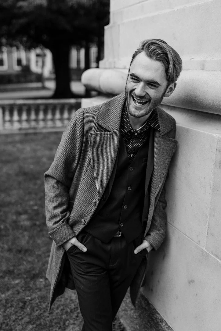 A man with a beard and styled hair laughing and leaning against a stone wall outdoors in a park, wearing a coat, button-up shirt, and pants.