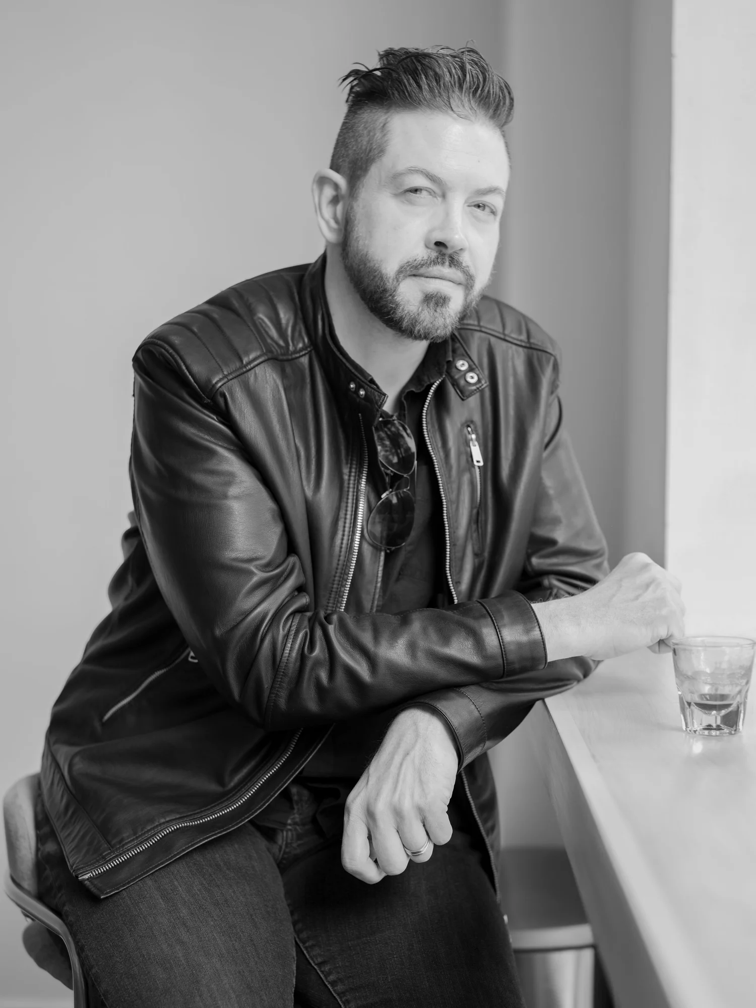 A man with a beard and styled hair sitting at a table, wearing a leather jacket and sunglasses hanging from his shirt, holding a glass on the table.