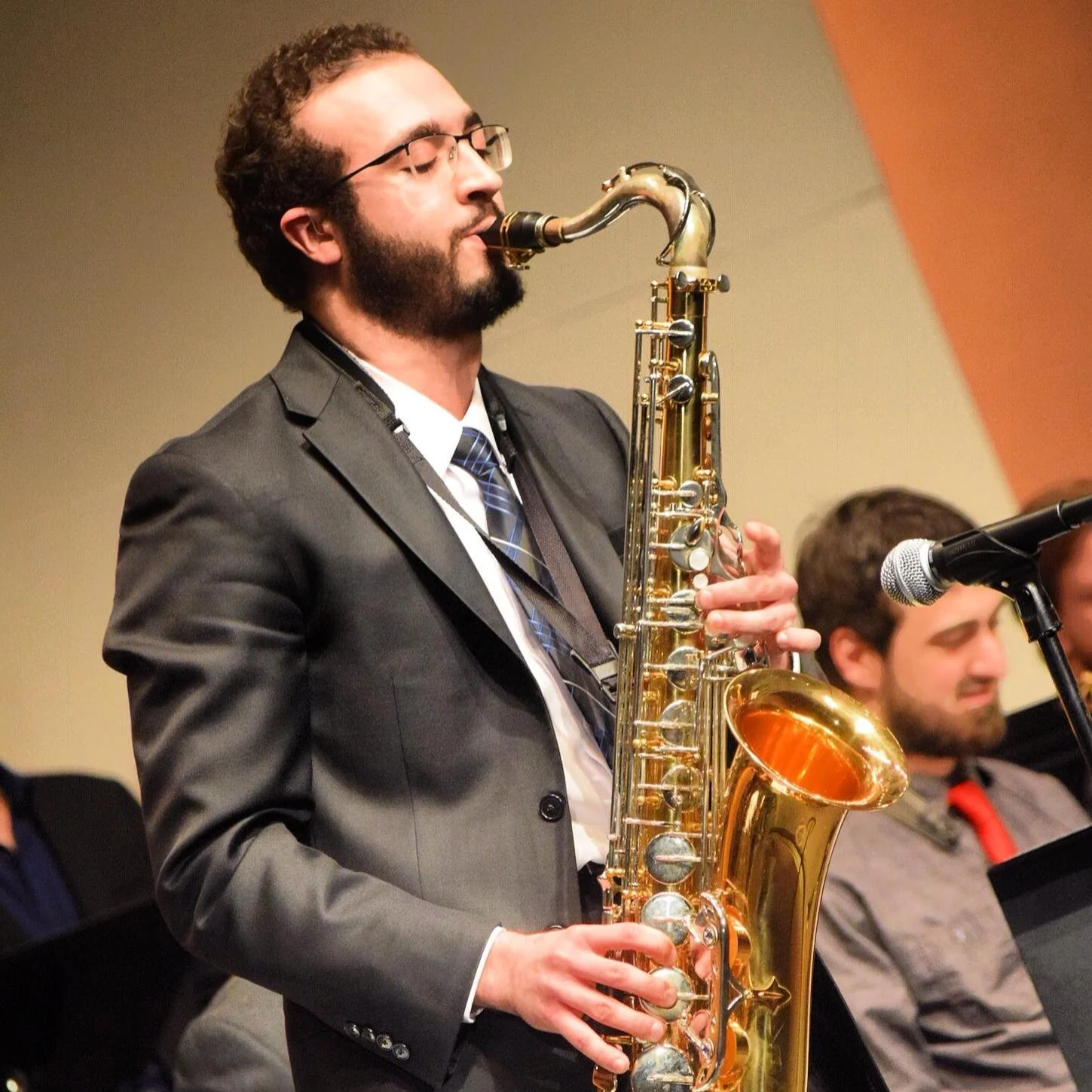 A man playing a saxophone during a performance, wearing glasses and a suit.