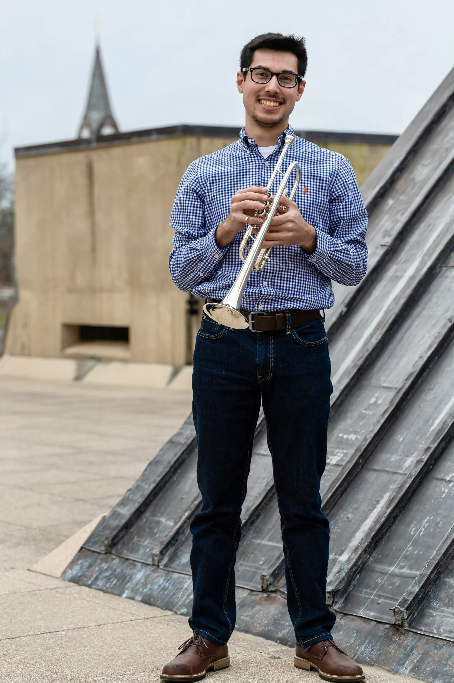 Young man with glasses, wearing a blue checkered shirt and jeans, holding a trumpet, standing on a rooftop with a chimney and building in the background.