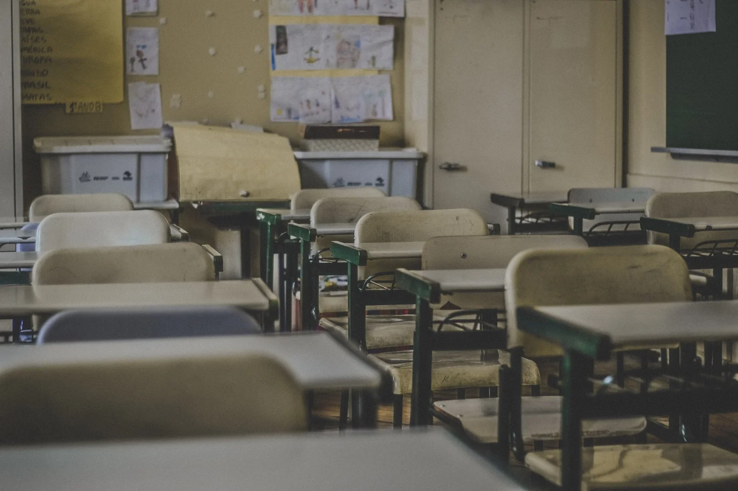 An empty classroom with beige desks and chairs arranged in rows, a bulletin board with children's drawings on the wall, and a chalkboard on the right.