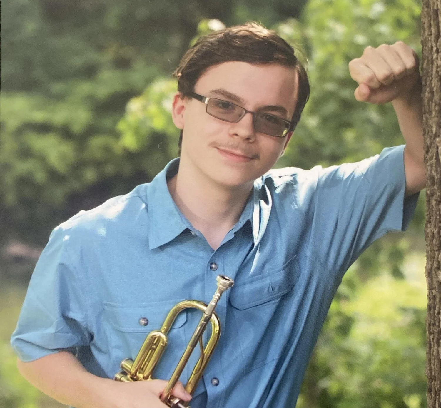 Young man with short brown hair, glasses, wearing a blue button-up shirt, smiling, outdoors in a wooded area, holding a trumpet, leaning with his right arm resting on a tree.