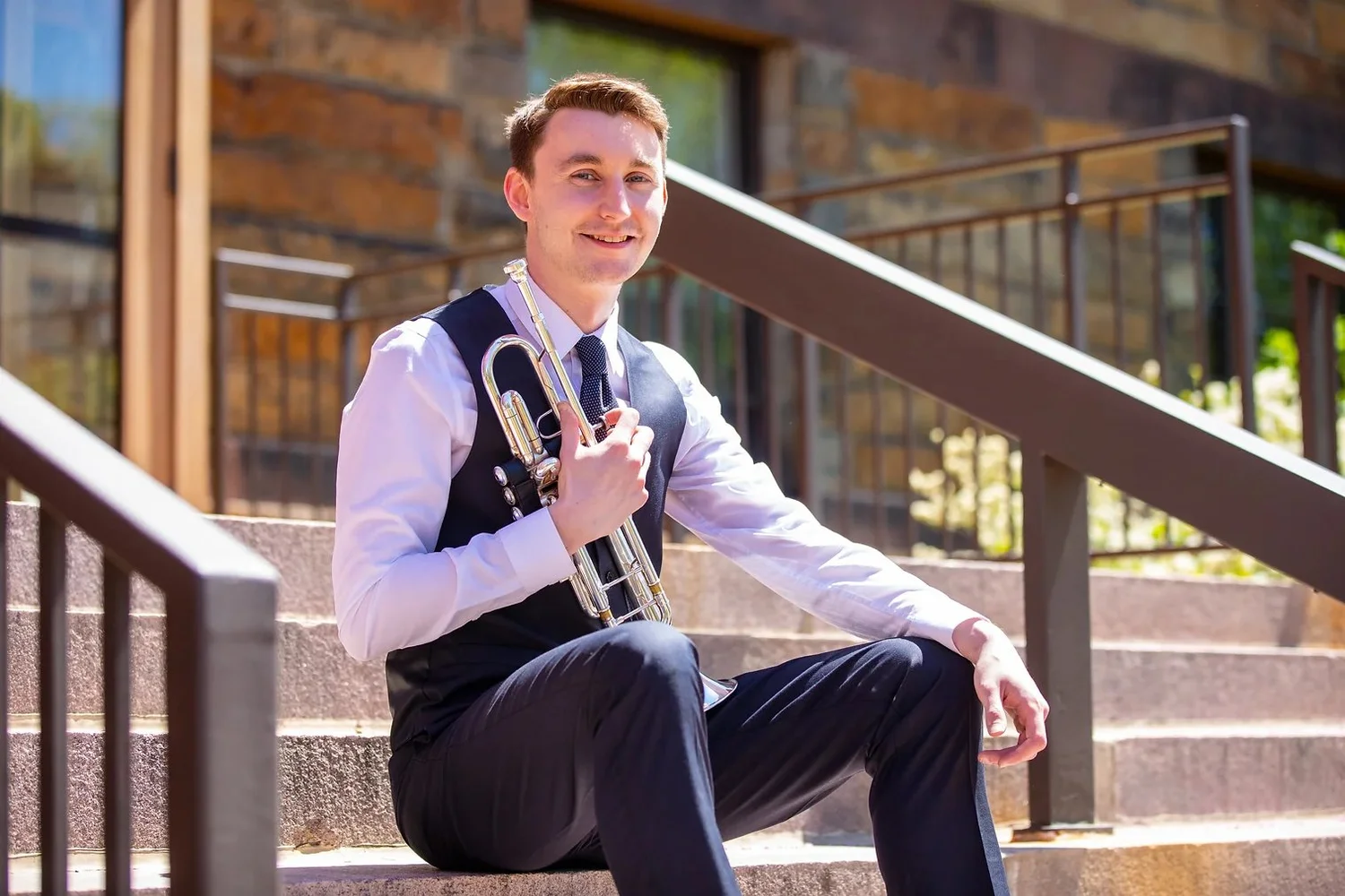 A young man dressed in a white shirt, dark vest, and tie, sitting on outdoor steps with a trumpet in hand, smiling at the camera.