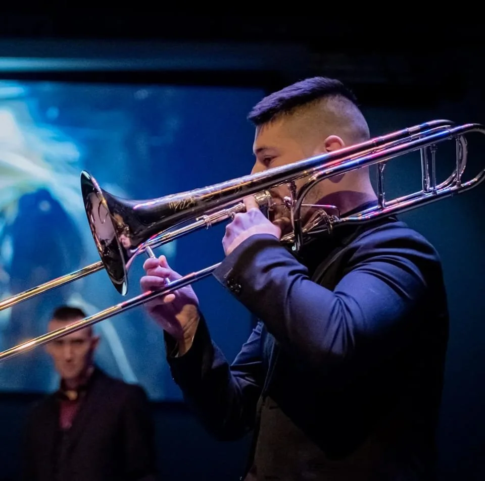 A man playing a trombone during a performance with a dark background and third person in the background.