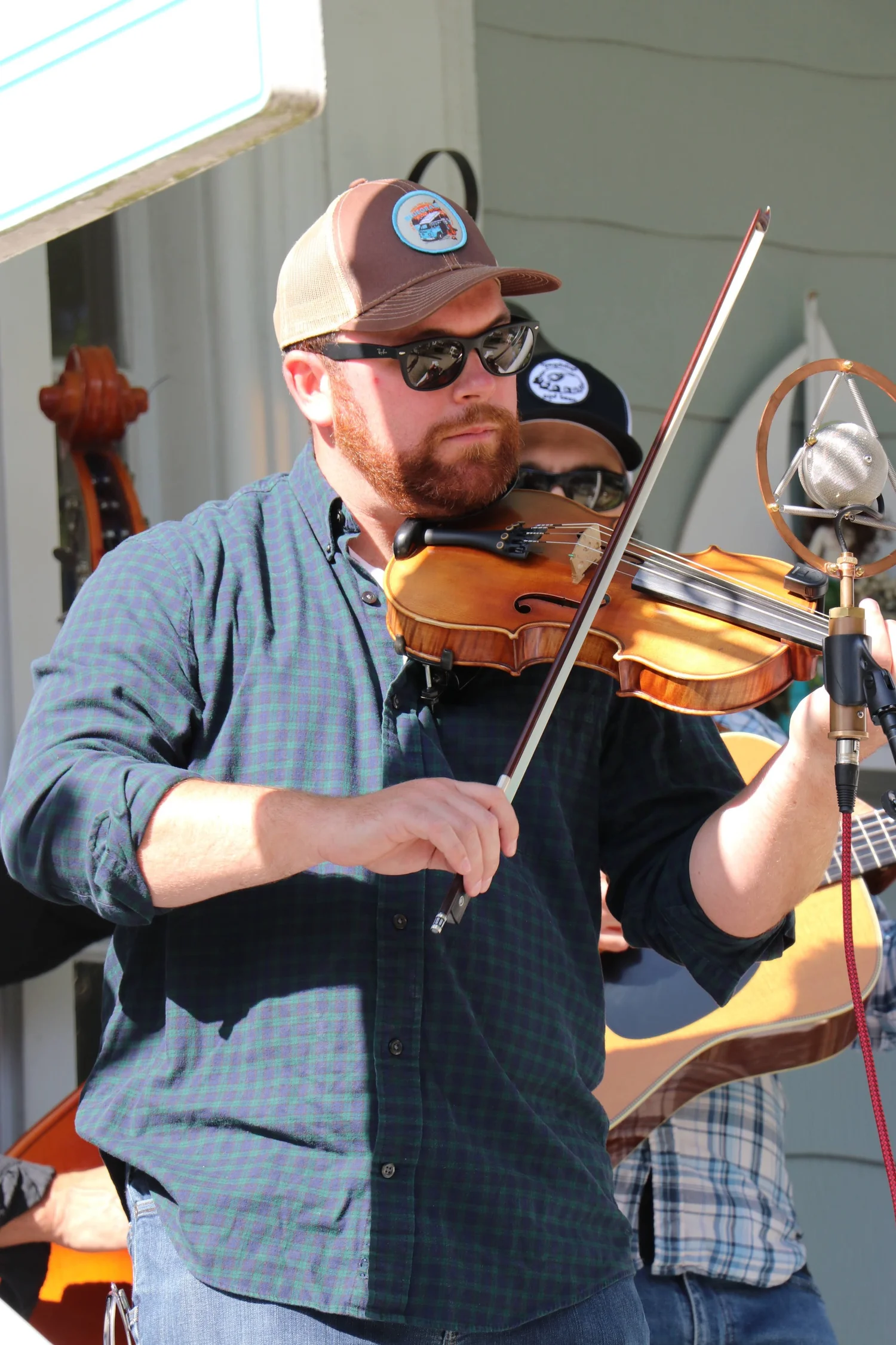 A man with a beard, wearing sunglasses and a plaid shirt, playing a violin outdoors.