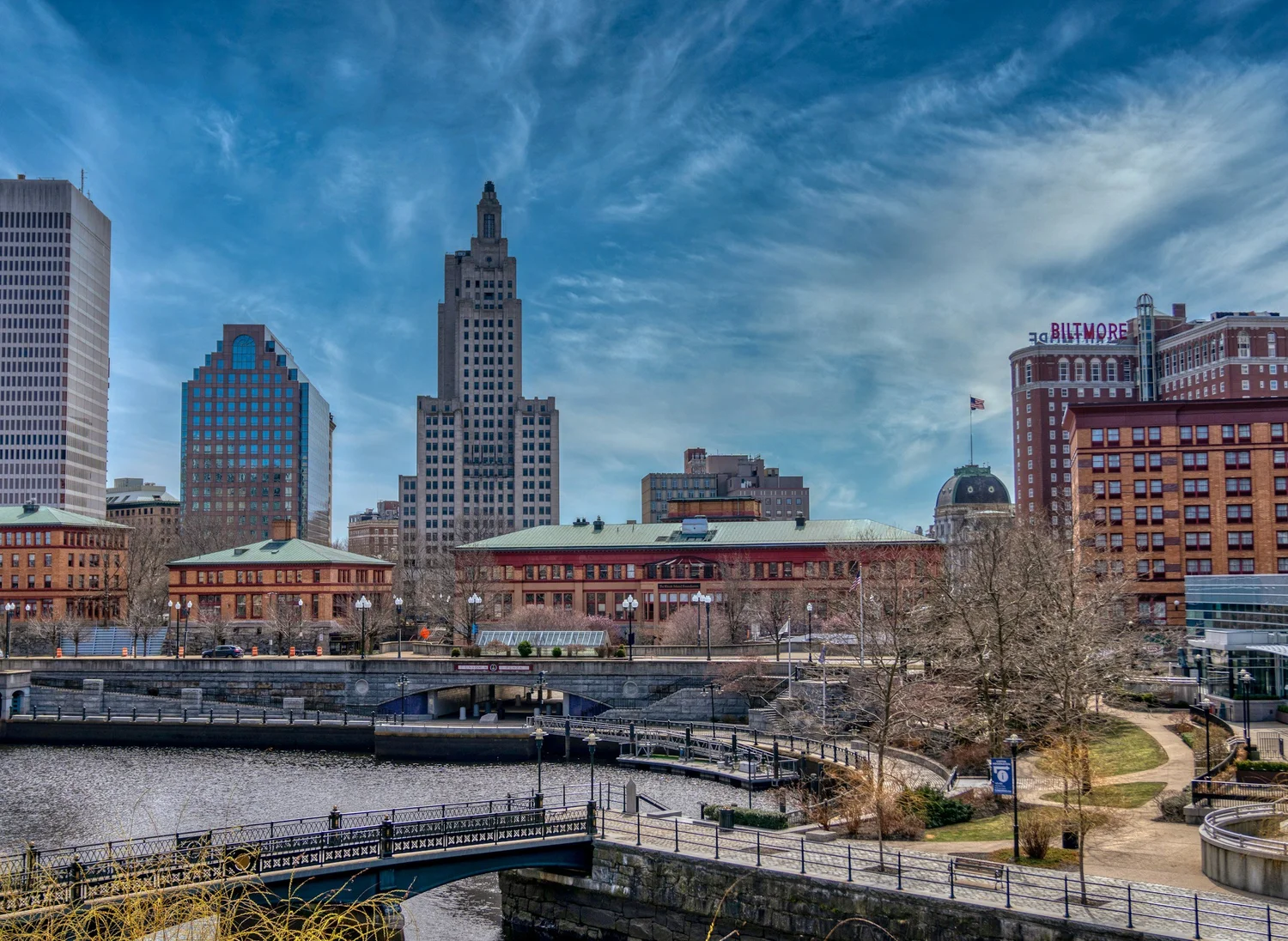 City skyline with tall buildings, a river, a park, and a bridge in the foreground on a clear day.