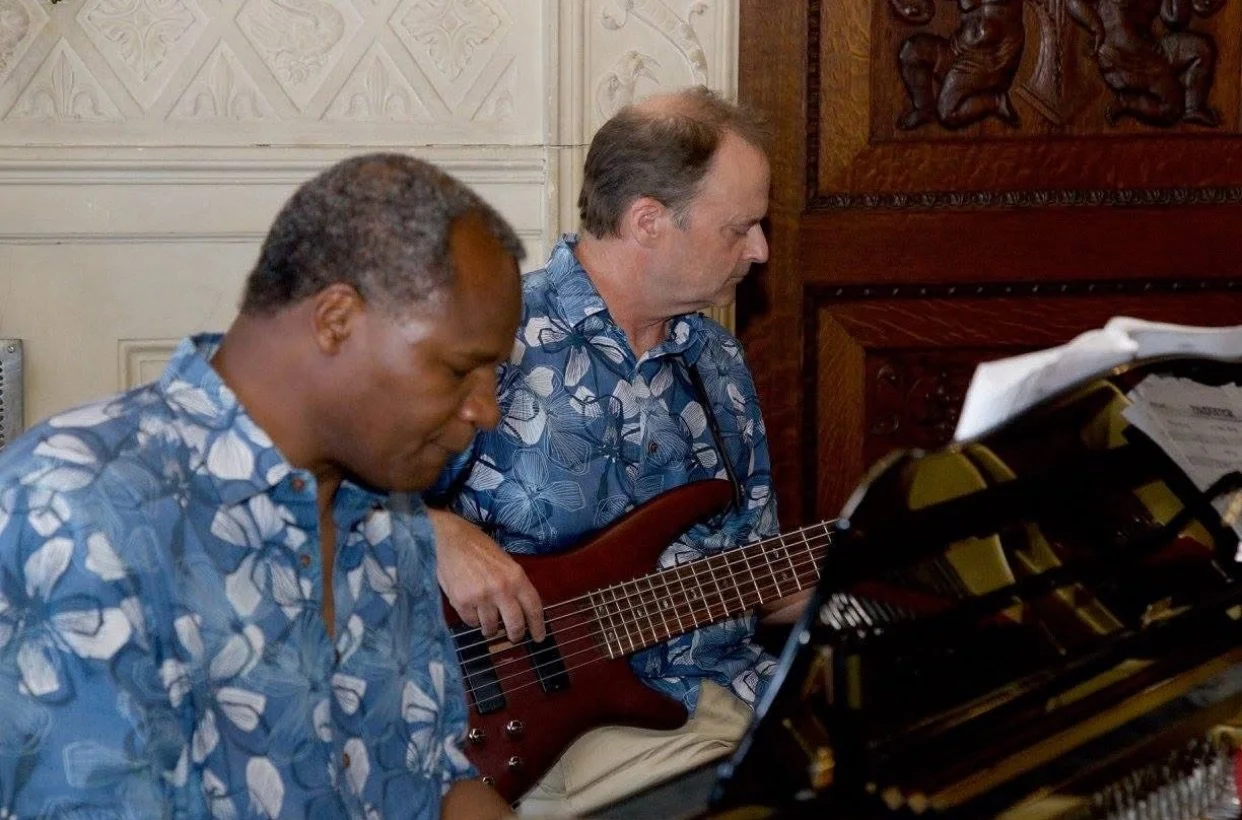 Two men wearing blue Hawaiian shirts sit and play instruments, one at a piano and the other with an electric bass guitar, in a room with ornate wall decor.