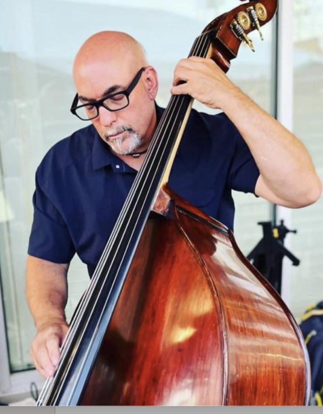 A man with glasses and a goatee playing a double bass, standing indoors.