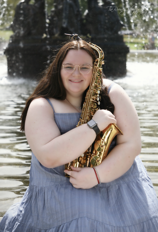 A young woman with long brown hair, wearing glasses, a blue dress, and a black smartwatch, smiling and holding a gold saxophone, sitting by a water fountain.