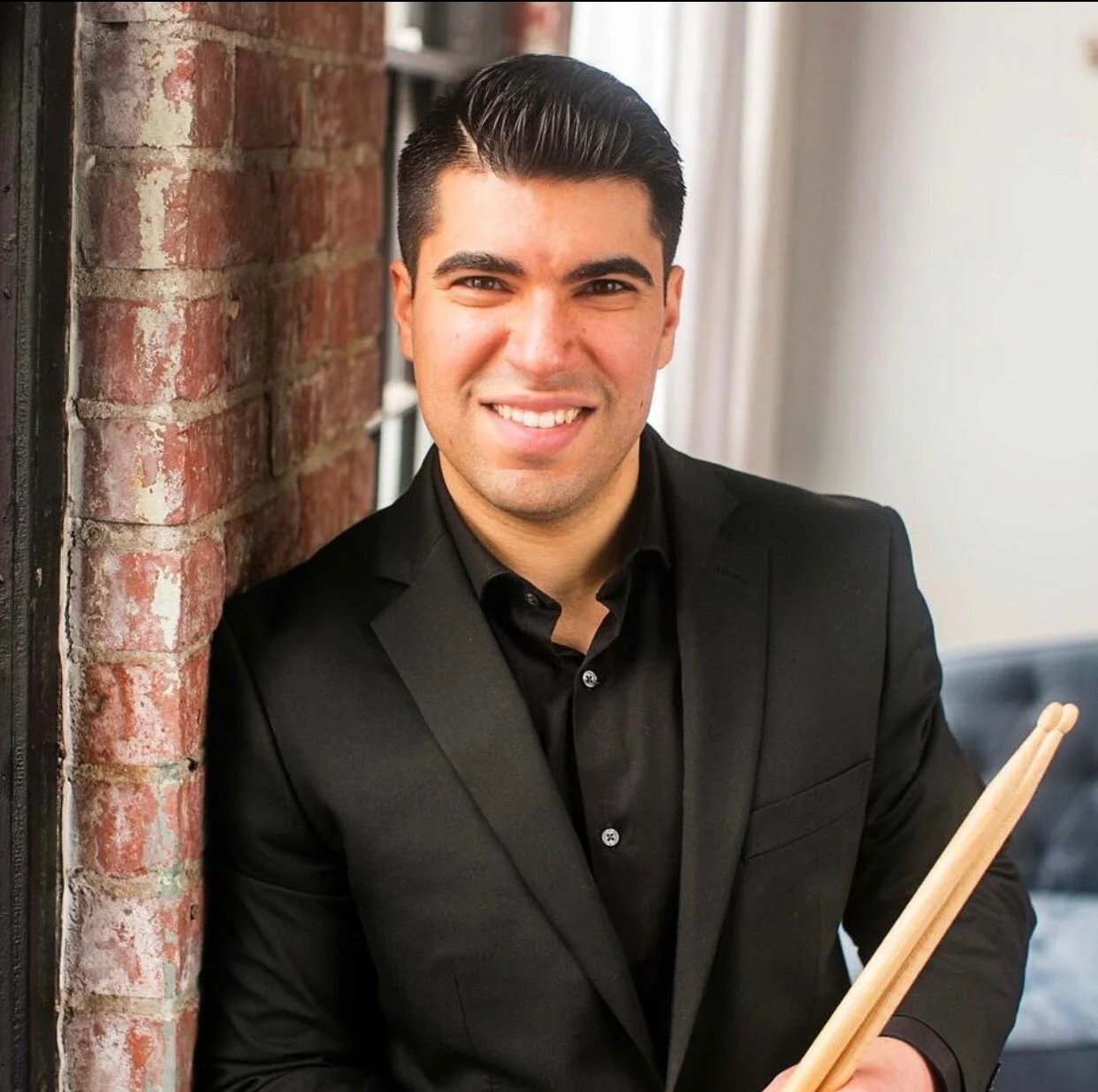 A young man smiling, wearing a black suit and shirt, holding drumsticks, leaning against a brick wall.