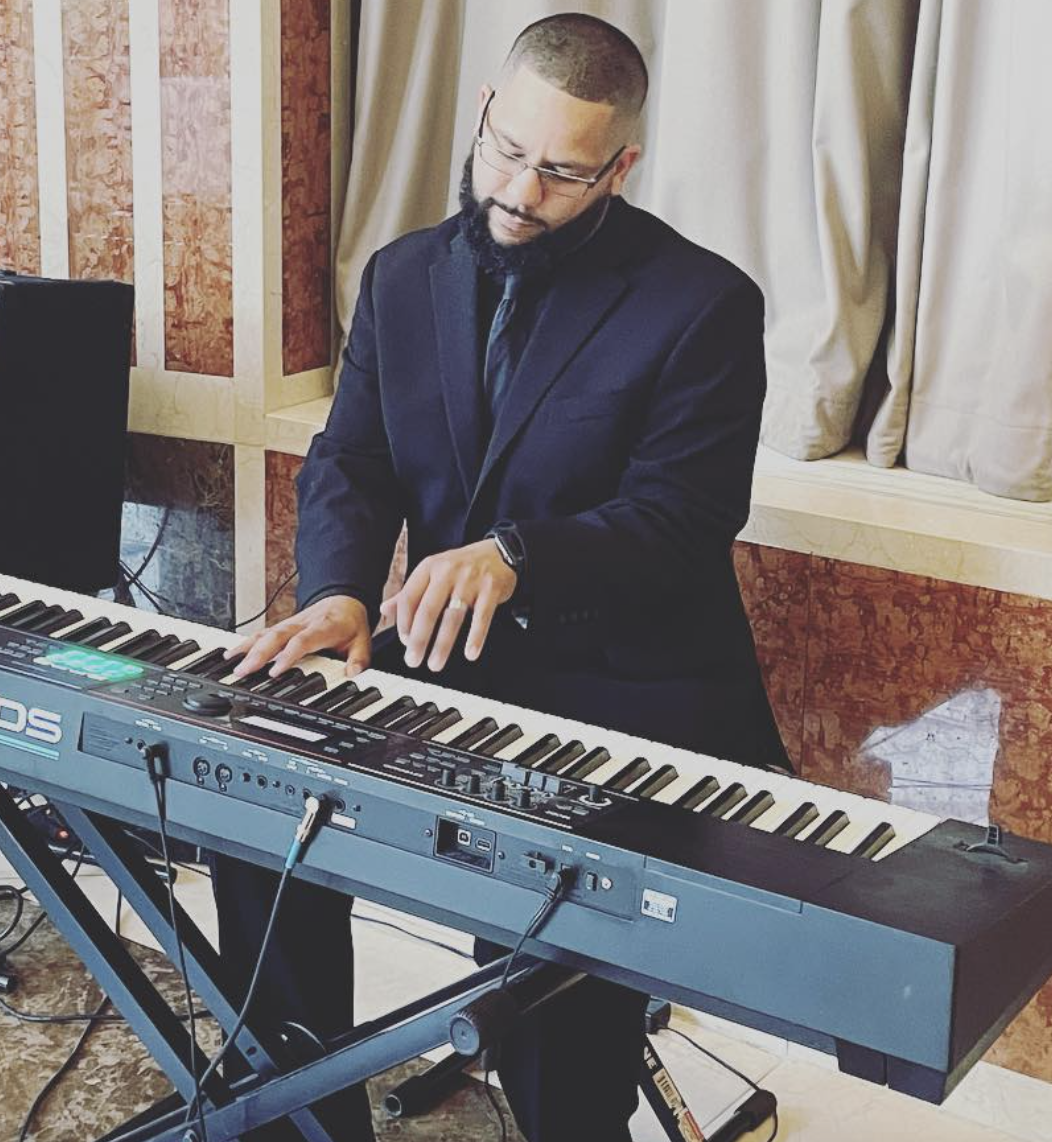 A man in a dark suit playing an electronic keyboard, with a violin ear monitor and glasses, in a room with patterned wood paneling and cream curtains.