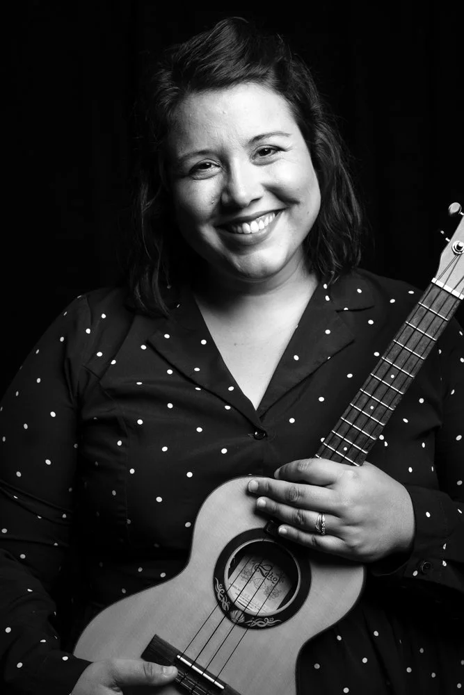 A woman smiling and holding a ukulele, poses in front of a dark background, wearing a polka dot blouse.