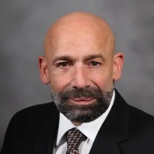 Professional headshot of a bald man with a beard wearing a suit and tie, against a gray background.