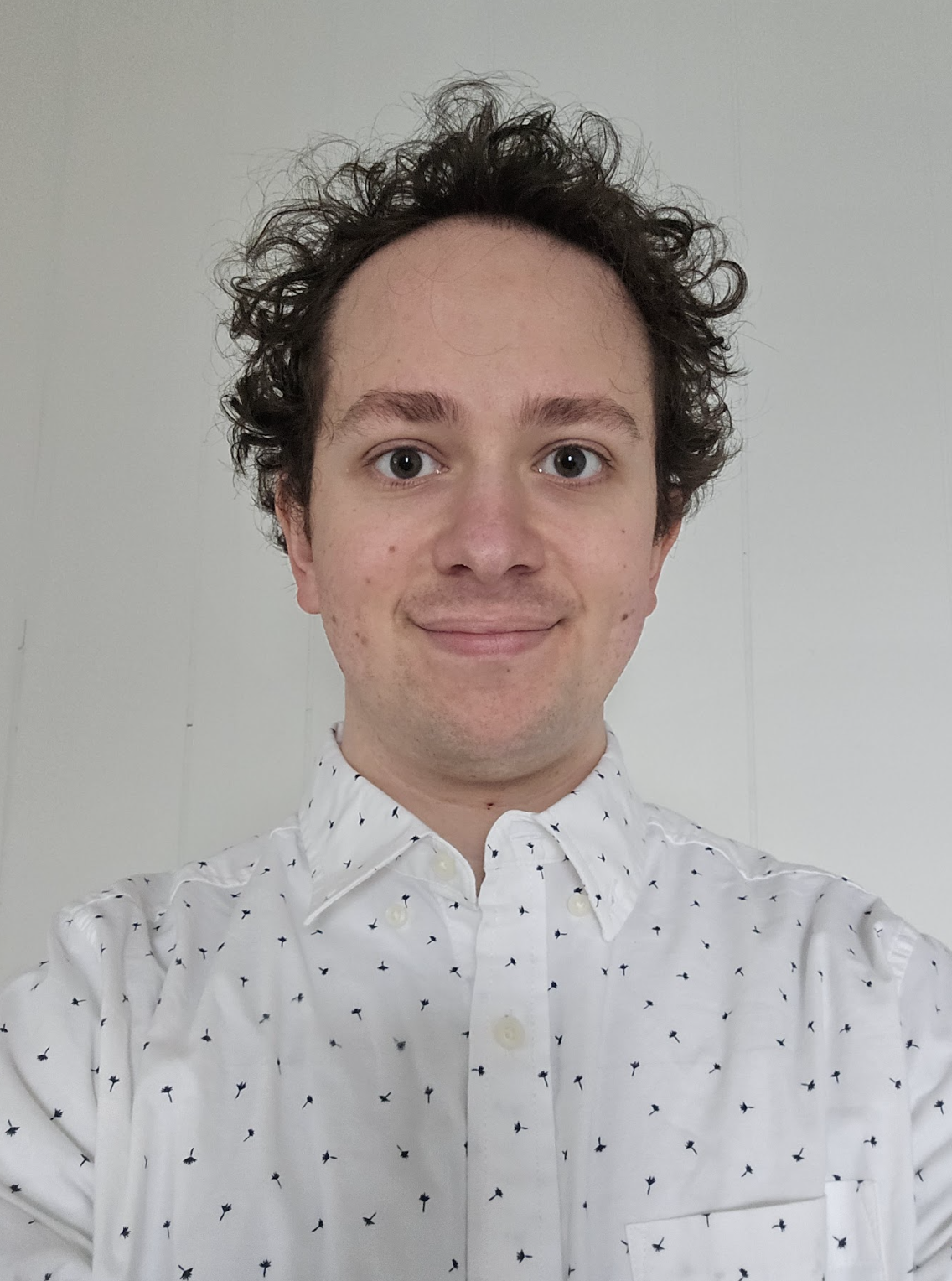 A young man with curly hair smiling in front of a plain white wall, wearing a white button-up shirt with small black patterns.
