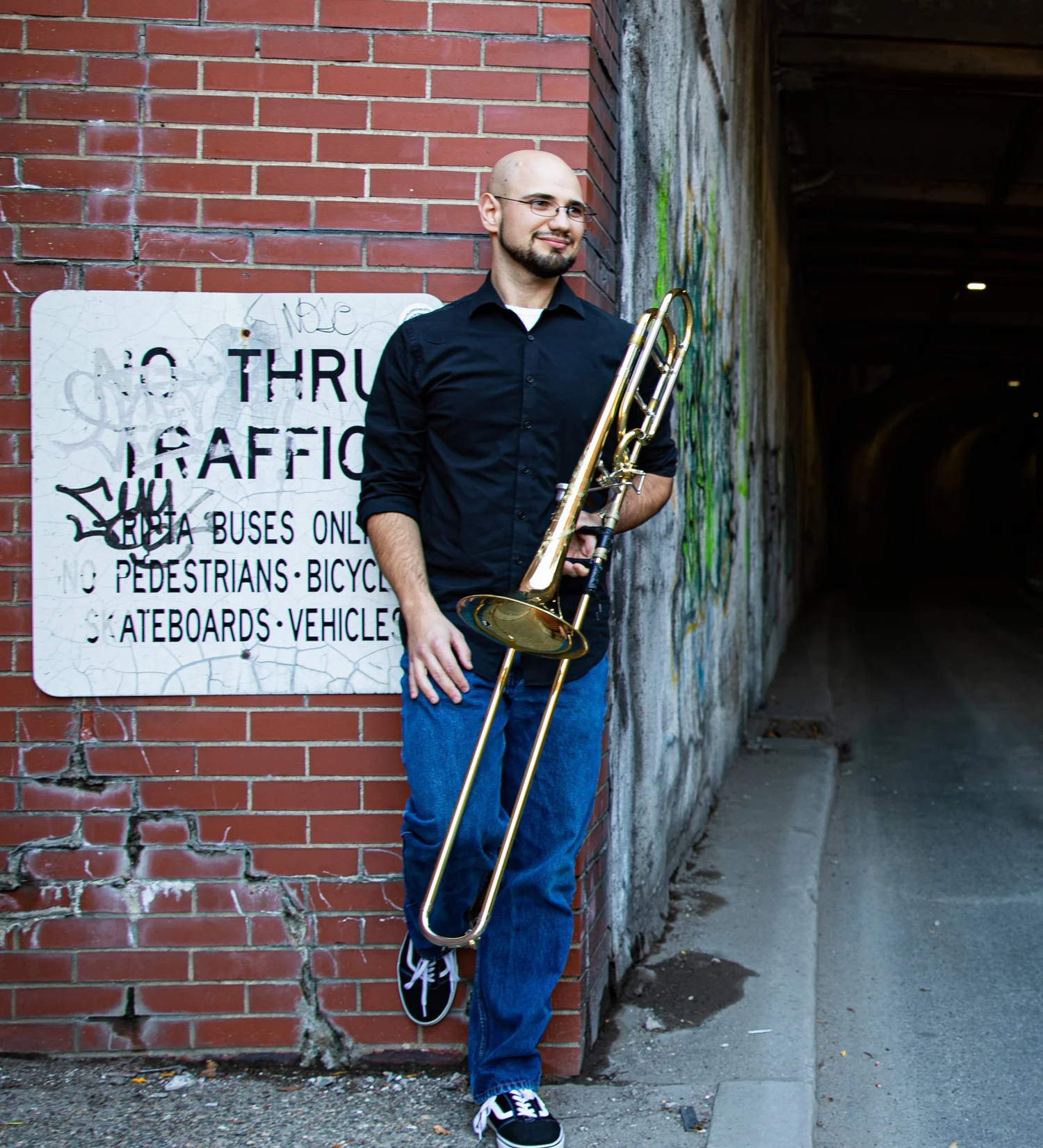 A man with glasses, a black shirt, and blue jeans, holding a trombone, standing near a brick wall and a graffiti-covered wall in an alleyway.