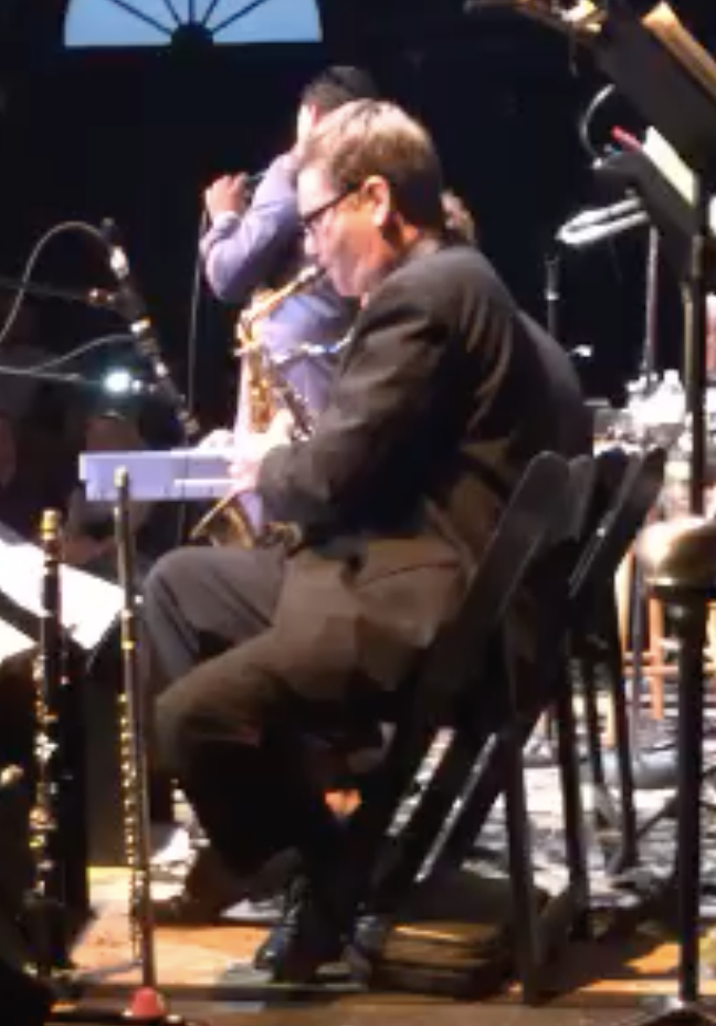 A man in a black suit playing a keyboard on stage with a saxophone, while a child wearing a gray shirt and glasses stands behind him, possibly holding or adjusting musical equipment, in a dimly lit setting.