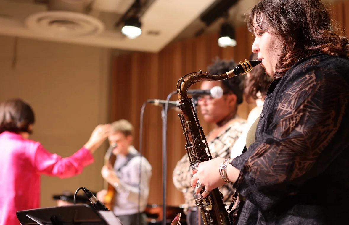 A woman playing the saxophone on stage during a band performance with other musicians and a woman conducting in the background.