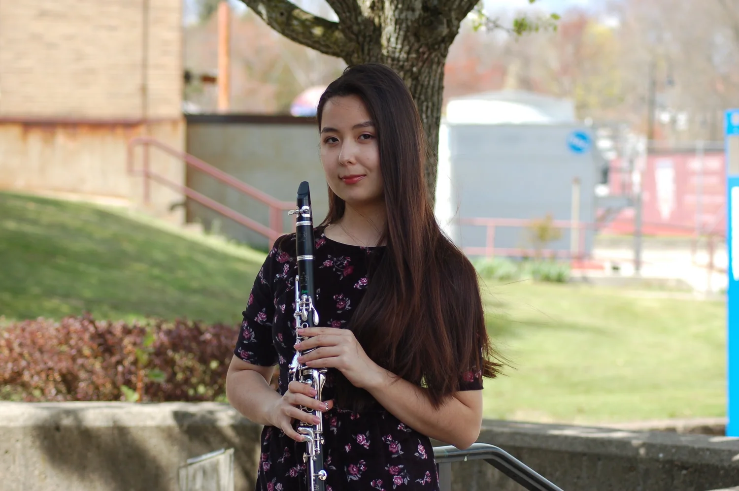 Young woman with long dark hair holding a clarinet outdoors, standing near a tree with a park or garden in the background.