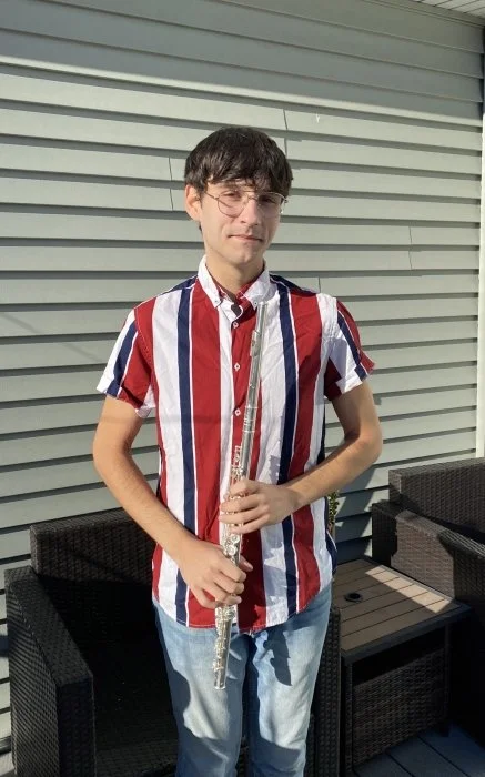 Young man standing outdoors on a patio, holding a flute, wearing glasses and a vertically striped red, white, and blue short-sleeved shirt with light blue jeans.