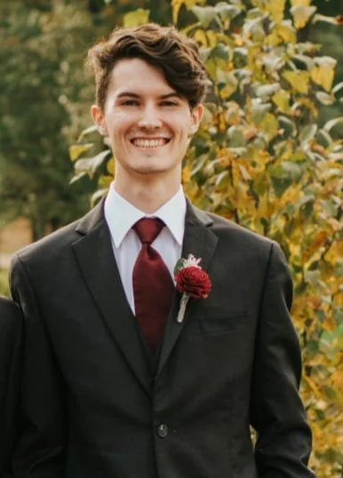 Young man in a black suit, white shirt, and red tie, smiling outdoors with autumn leaves in the background.