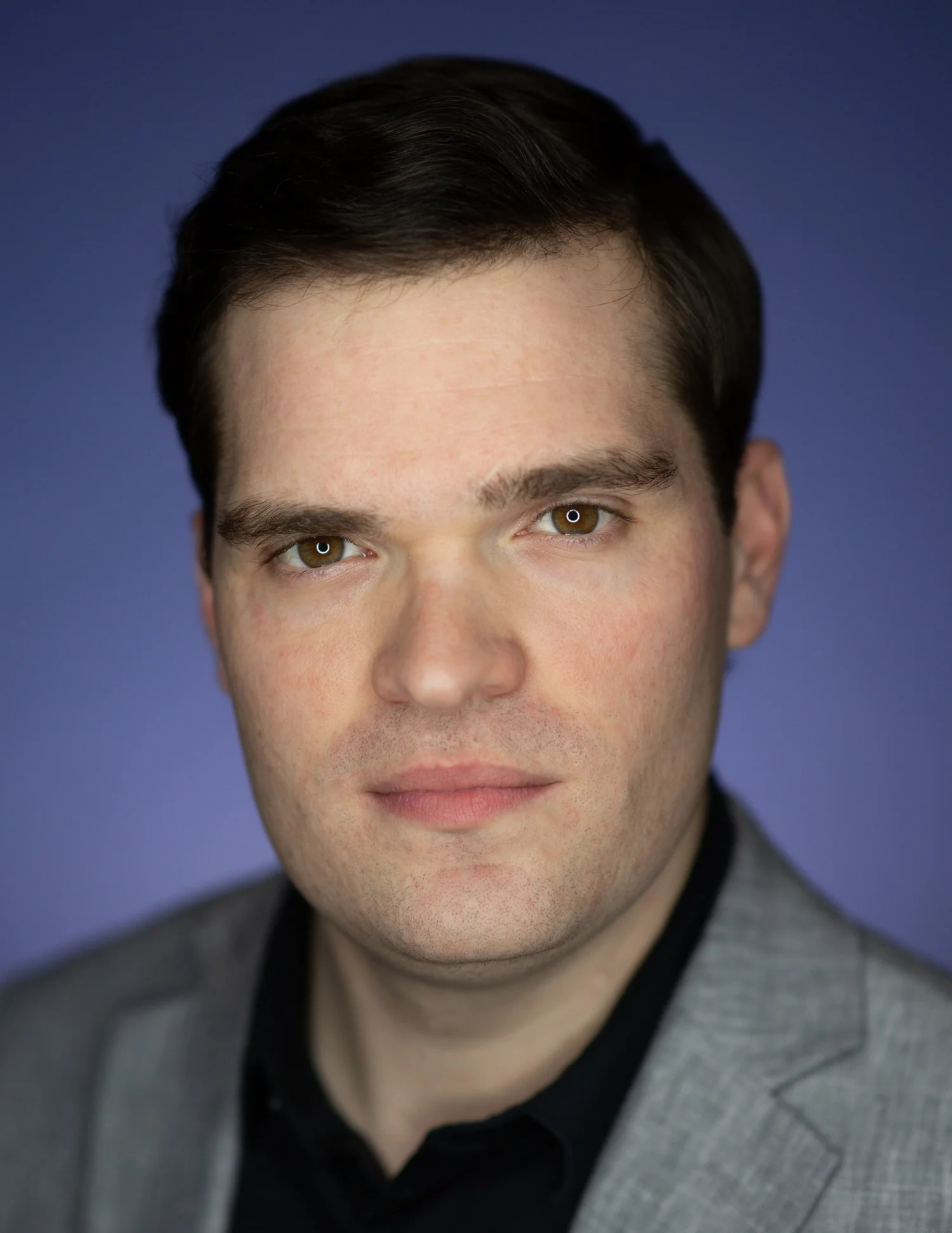 Headshot of a man with dark hair, fair skin, wearing a gray blazer and black shirt, against a blue background.