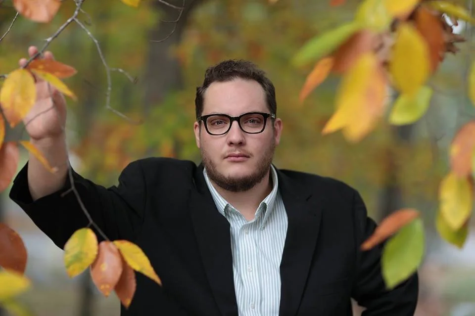 A man wearing glasses and a black blazer standing outdoors among autumn trees with yellow and orange leaves.