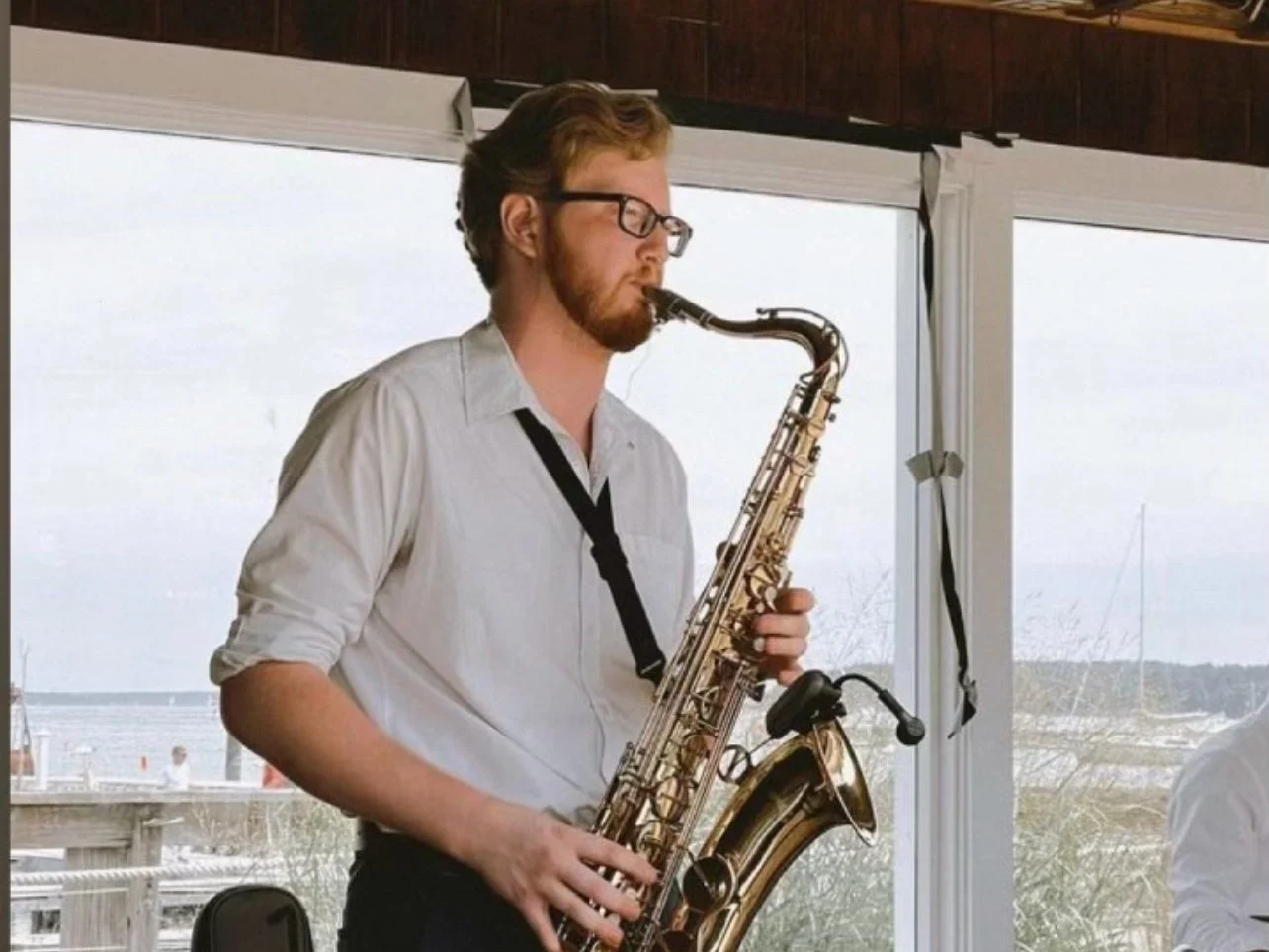 A man with glasses and a white shirt playing a saxophone indoors near large windows with a dock and boats outside.