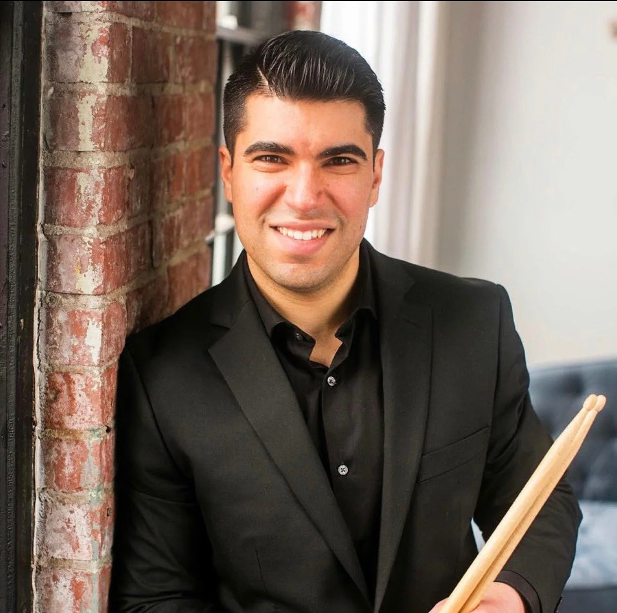 A man in a black suit smiling, holding a wooden drumstick, leaning against a brick wall.