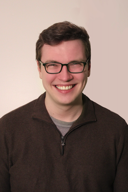 A young man with glasses smiling, wearing a dark zip-up sweater over a gray shirt, standing against a neutral background.
