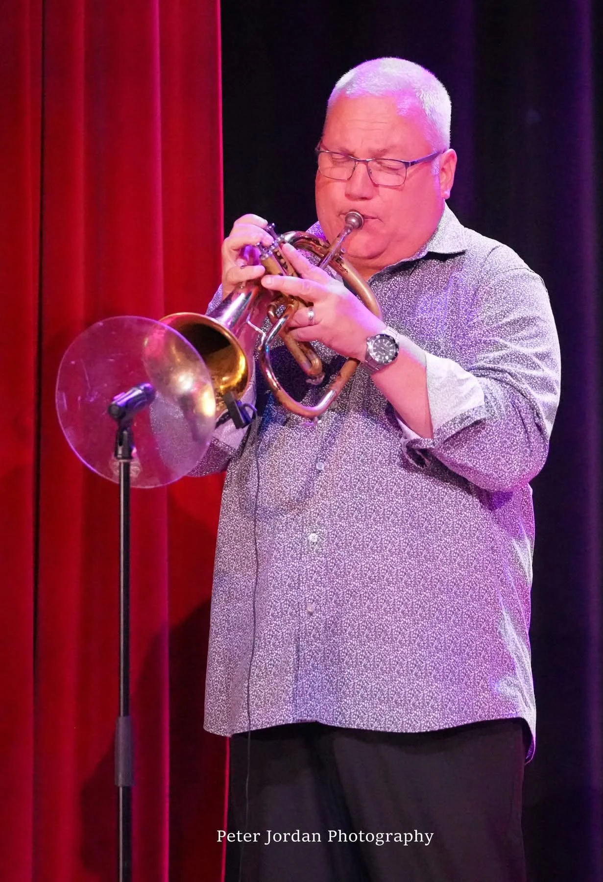A man with glasses and a short haircut playing a trumpet on stage with red curtains in the background.