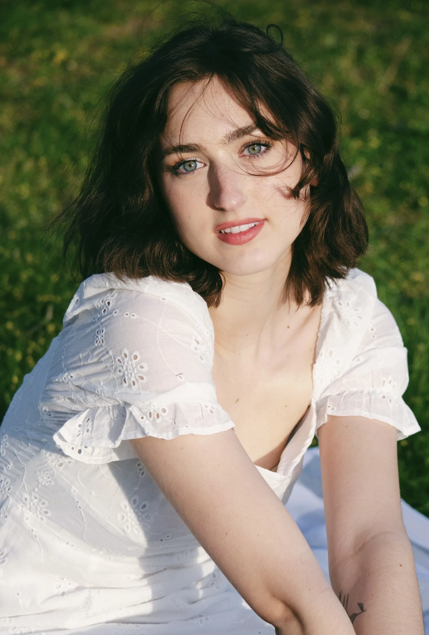 A young woman with dark, wavy hair and blue eyes sitting outdoors on green grass, wearing a white eyelet dress, smiling softly at the camera.