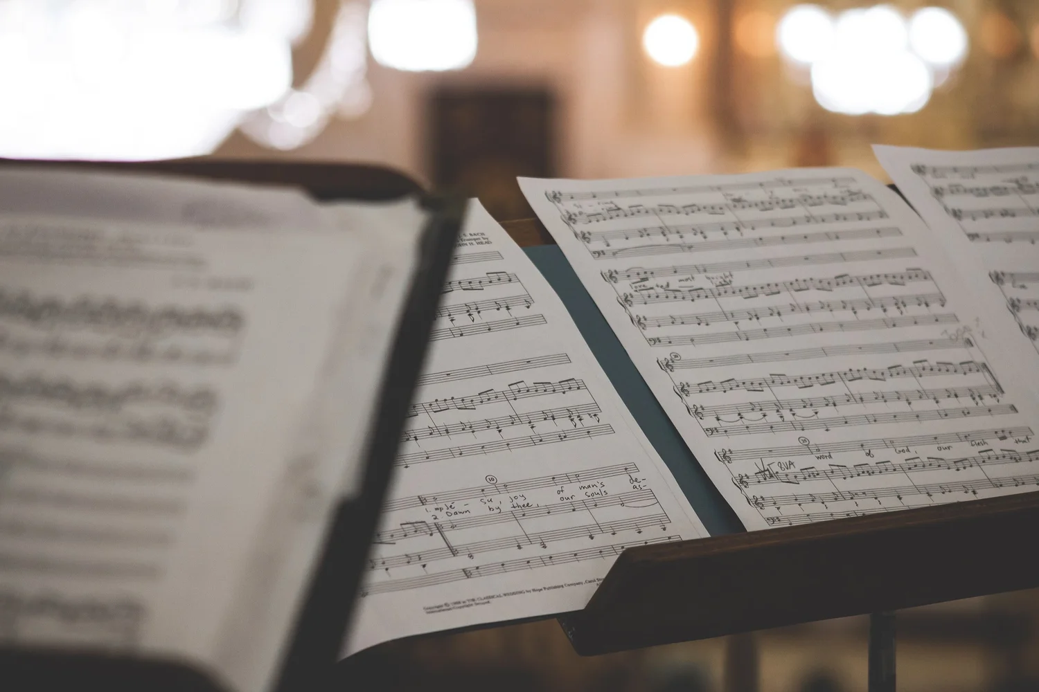 Open music sheet books on a stand with other music sheets, in a dimly lit setting with blurred background.