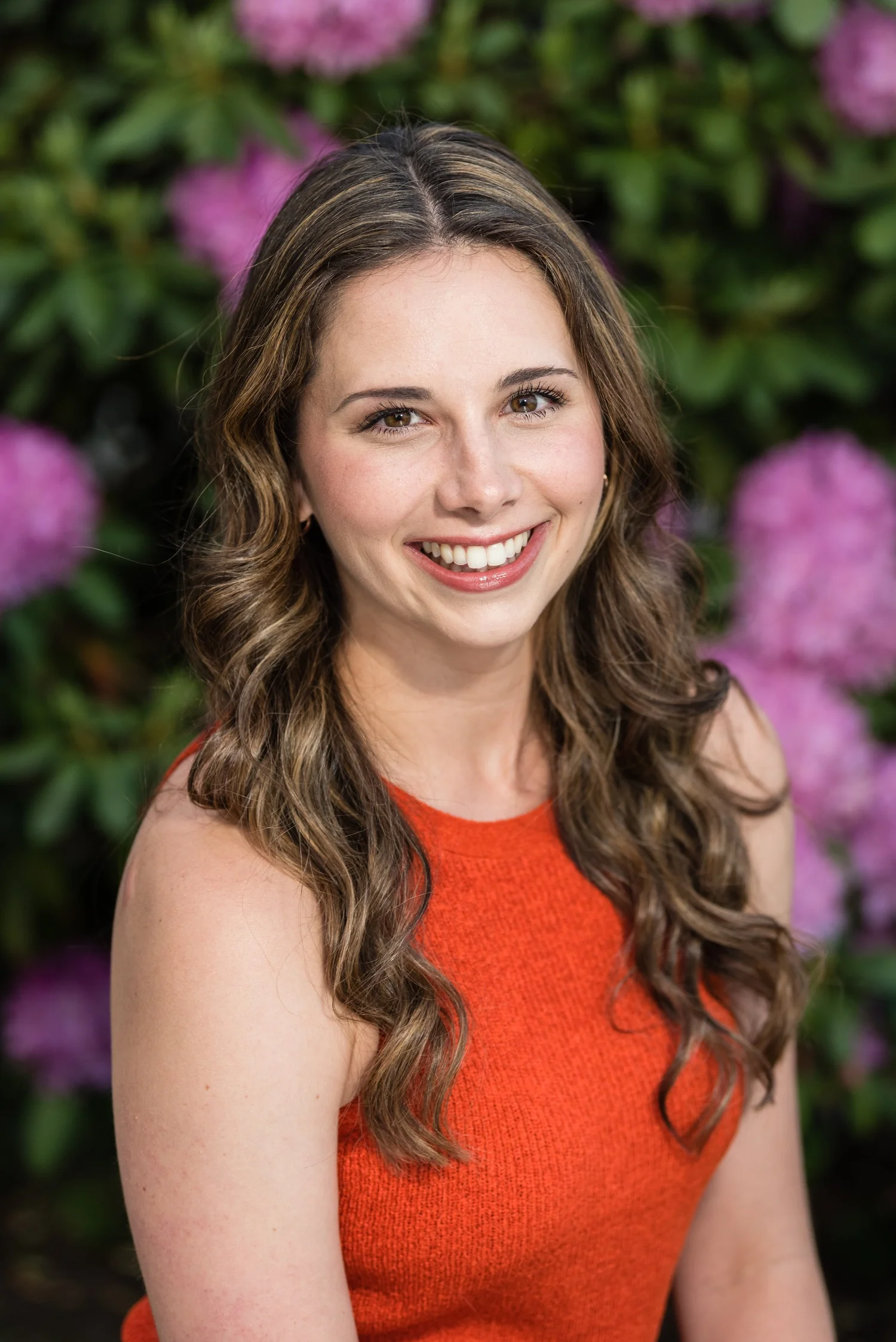 A smiling woman with long, wavy brown hair wearing an orange sleeveless top, standing outdoors with pink flowers and green foliage in the background.