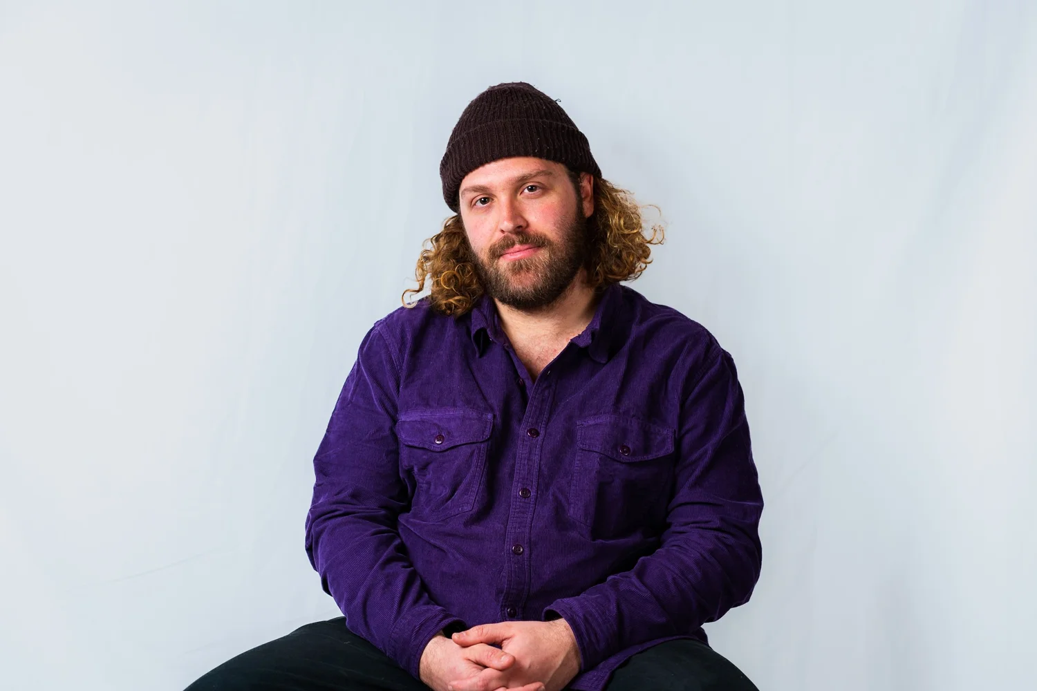 A man with curly hair and a beard, wearing a brown knit cap and purple shirt, sitting against a light background.