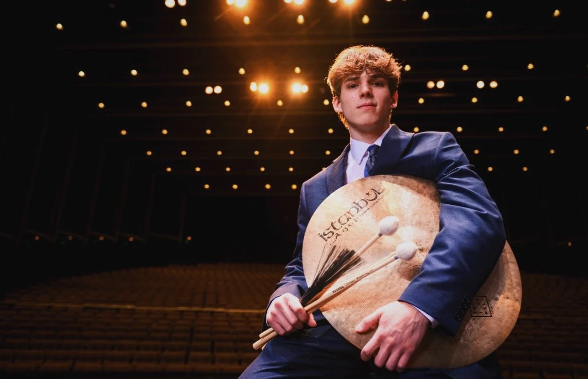 Young man in suit holding cymbals and drumsticks on stage with bright lights overhead in an empty theater.