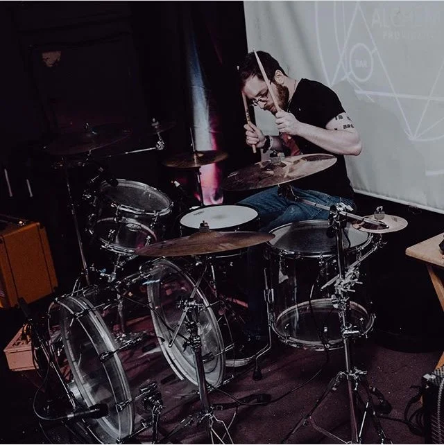 Man playing drum set indoors with a projection screen in the background.