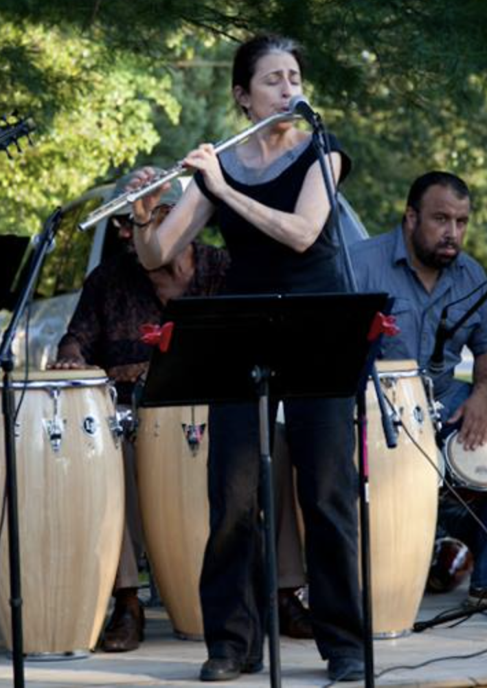 Woman playing flute in an outdoor band with percussion instruments, two men playing drums behind her, trees and green foliage in the background.