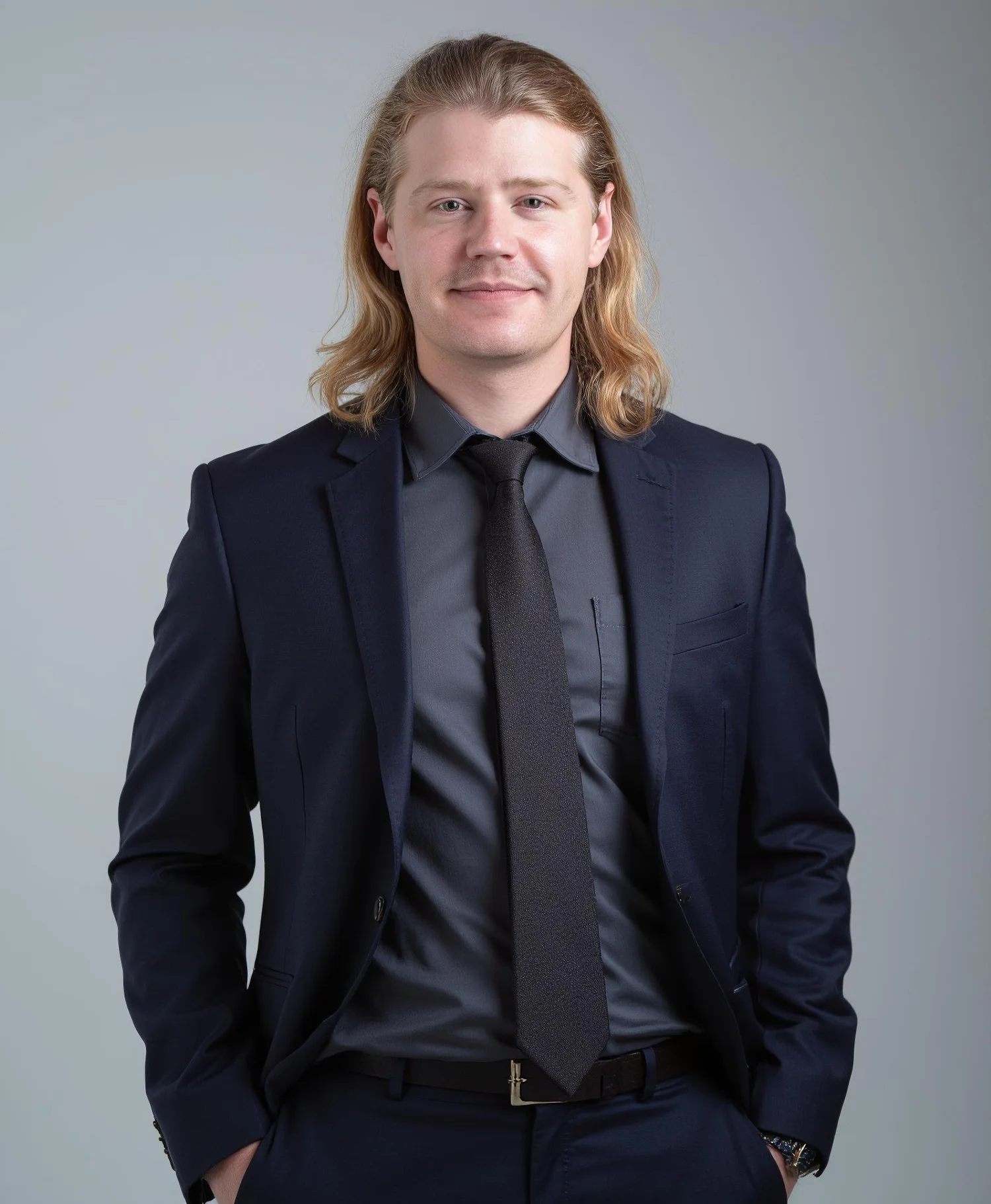 A young man with long blonde hair, wearing a dark suit, gray shirt, and black tie, standing with hands in pockets against a neutral gray background.