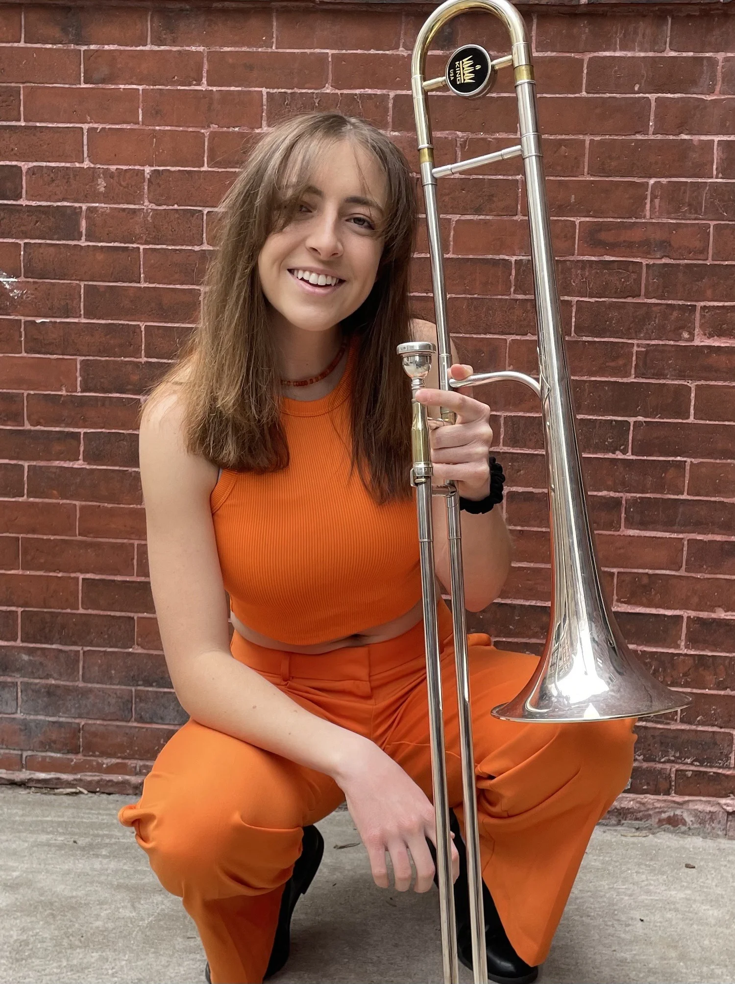 Woman in an orange sleeveless top and matching pants holding a trombone in front of a red brick wall. She is smiling and kneeling down on one knee.