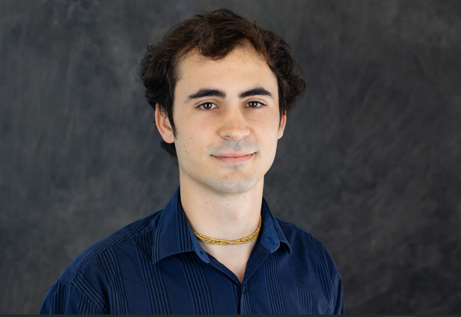 A young man with dark, curly hair and fair skin wearing a dark blue shirt with vertical stripes and a gold beaded necklace, standing in front of a dark, textured background.