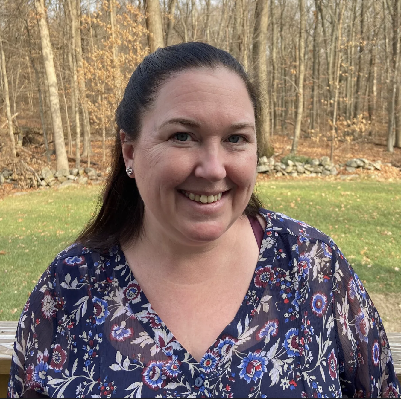 A woman with dark brown hair in a ponytail, wearing a floral navy blue blouse, smiling outdoors on a wooden deck with a grassy yard, trees, and a stone wall in the background during autumn.