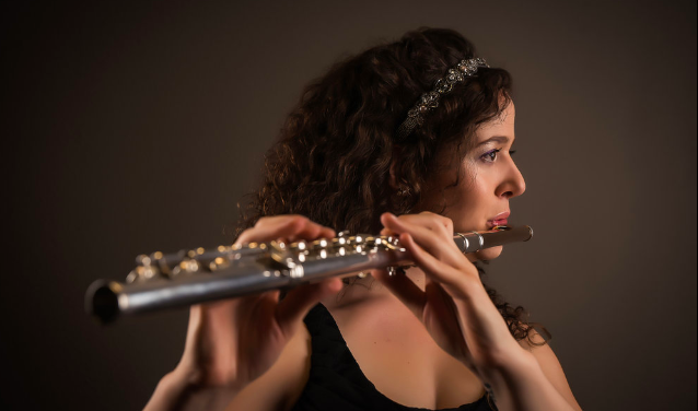 A woman with curly brown hair and a decorative headband playing a flute against a neutral background.