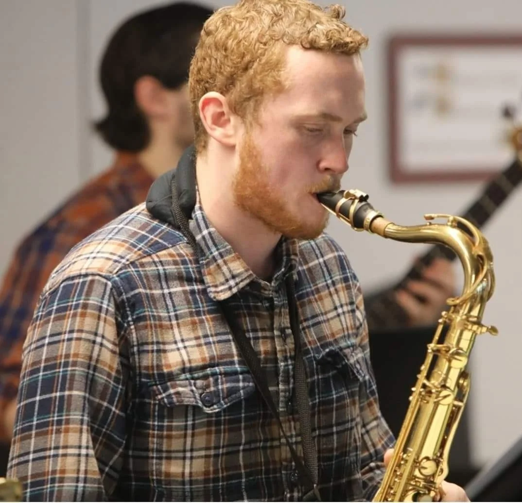 A young man with red hair and beard playing a gold saxophone, wearing a plaid shirt.