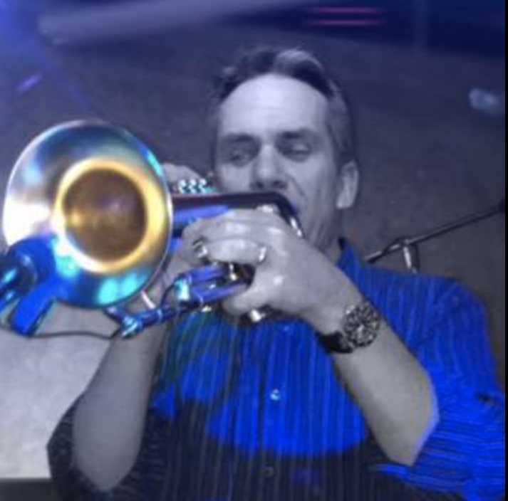 A man playing a trumpet in a dimly lit indoor setting, wearing a blue striped shirt and a watch.