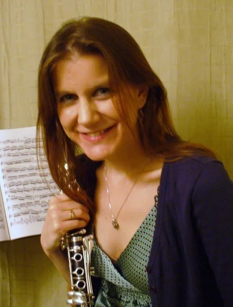 A woman with long reddish-brown hair smiling, holding a silver flute, standing next to a music stand with sheet music.