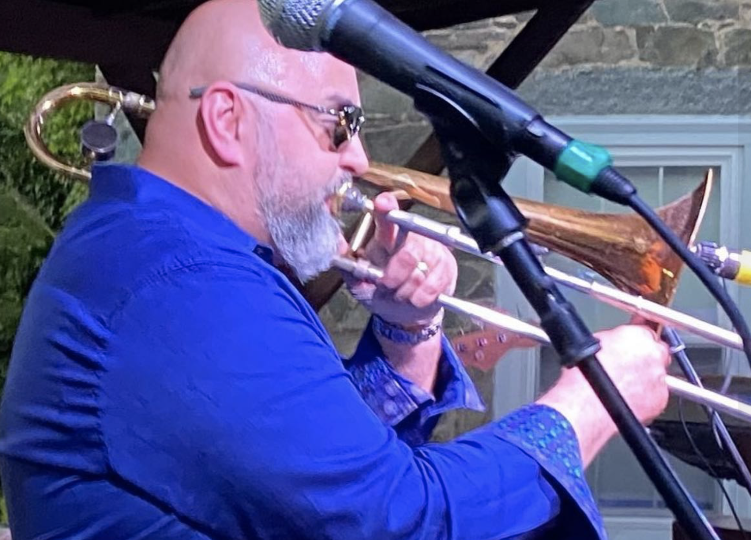 A man with a beard, sunglasses, and a blue shirt playing a trombone during a musical performance, with microphones set up in front of him.