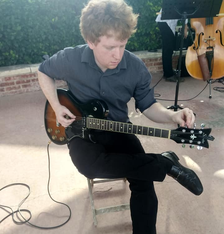 A young man with curly hair sitting on a chair, tuning a black electric guitar, with a cello and other musical equipment in the background.