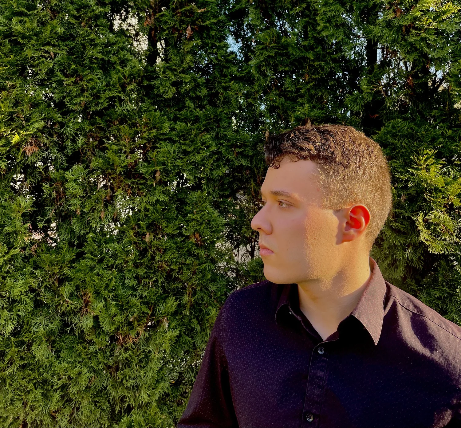 Side profile of a young man with short curly hair, wearing a black shirt, standing outdoors against a background of green dense bushes.
