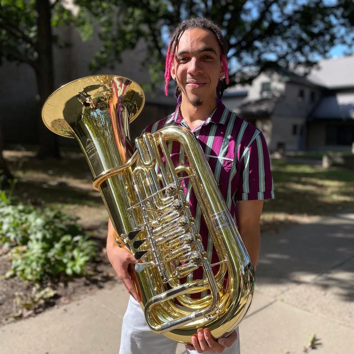 A young man with pink-tipped dreadlocks holds a shiny brass tuba outdoors on a sunny day, with trees and houses in the background.